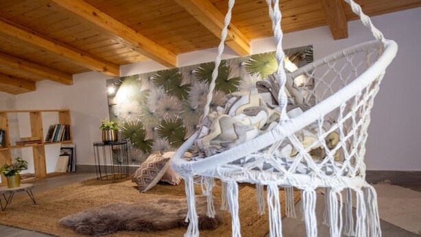 Una habitación con una silla colgante de macramé, techo de madera, una estantería y una pared decorada con plantas tropicales.