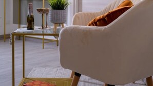 Una sala de estar moderna con un sillón blanco, un espejo con marco dorado, una mesita con una planta y una puerta al fondo.