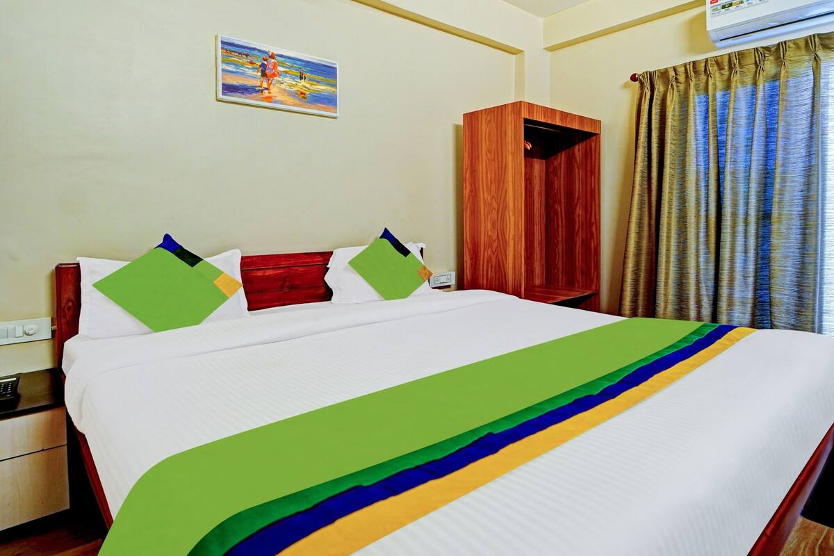 A hotel room with a bed, a nightstand, a wooden wardrobe, a painting on the wall, and a window with curtains.