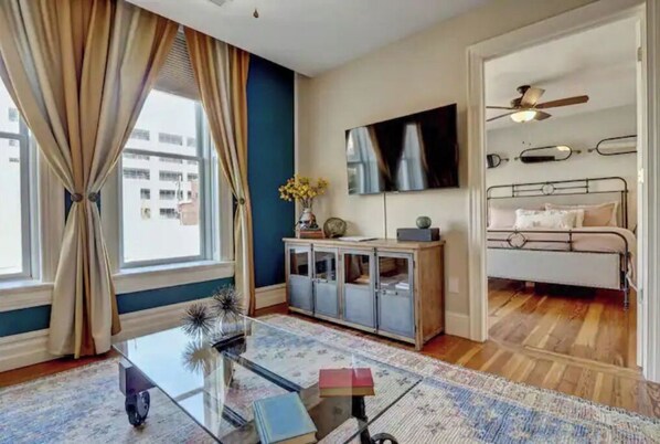 A modern hotel room with a large window, a flat-screen TV, a bed, a ceiling fan, and a glass coffee table.