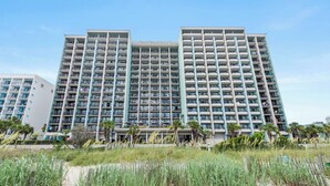 A high-rise hotel with multiple balconies, a sandy beachfront in the foreground, and palm trees. - Compass Cove Mariner Tower 624 - Ocean Front Condo - Pools, Lazy River, Hot Tub (Myrtle Beach)