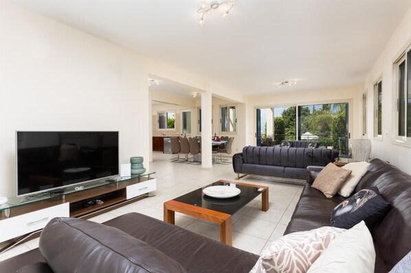 A modern living room with a flat-screen TV, leather sofas, a glass coffee table, and a dining area in the background.