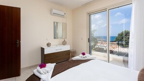 Un dormitorio con cama, cómoda, espejo y puerta corredera de vidrio que da a un balcón con vista al mar.