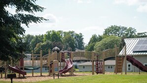 Un terrain de jeux avec des toboggans et des structures d’escalade, entouré d’arbres et avec une maison en arrière-plan.