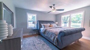 Une chambre dotée d’un grand lit, d’un ventilateur de plafond et de deux fenêtres donnant sur un paysage verdoyant.
