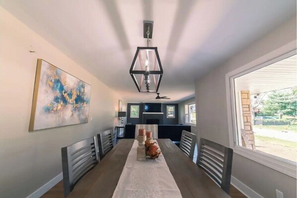 A modern dining area with a long table, benches, a hexagonal pendant light, and a large abstract painting on the wall.