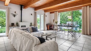 A spacious living area with a high wooden ceiling, a large bed with patterned bedding, a dining table with chairs, and a view of greenery outside.