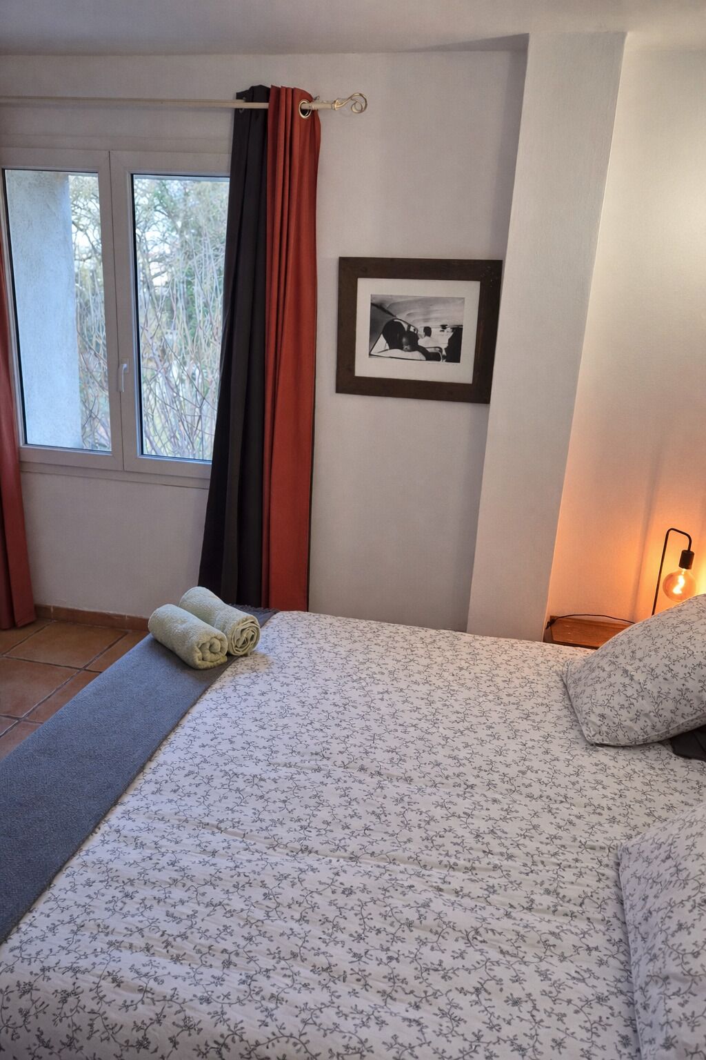 Une chambre à coucher avec un lit, une fenêtre avec des rideaux, un tableau encadré au mur et une lampe de chevet.