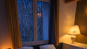 A bedroom with a bed, pillows, a bedside lamp, a window with curtains, and a view of trees outside.