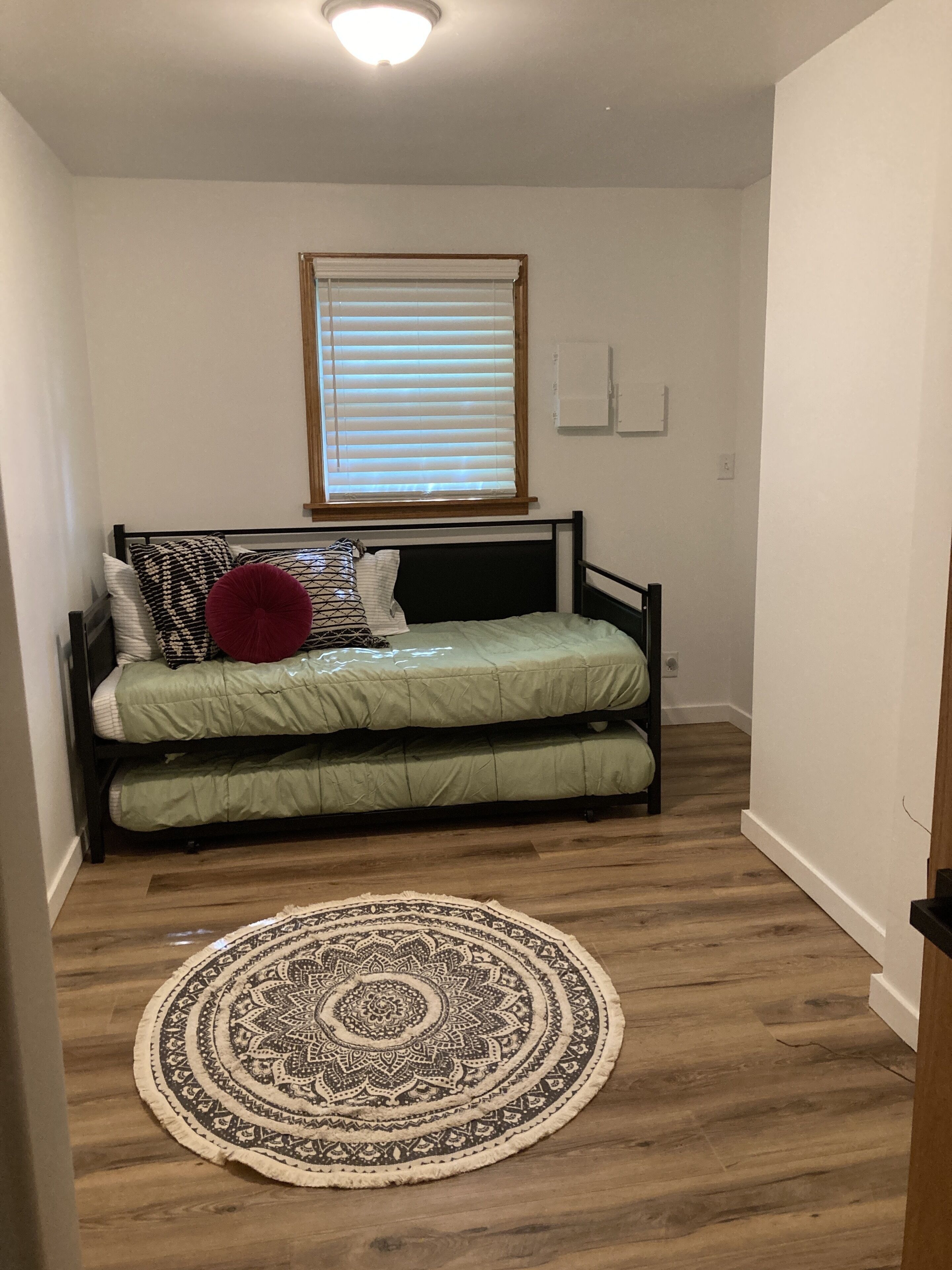 A small, single-bed room with a wooden floor, a round patterned rug, and a window with blinds.