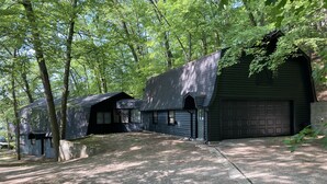 A rustic cabin with a large garage, surrounded by dense trees. - Lakefront Cottage with Private Beach, Sleeps 14 (Newaygo)