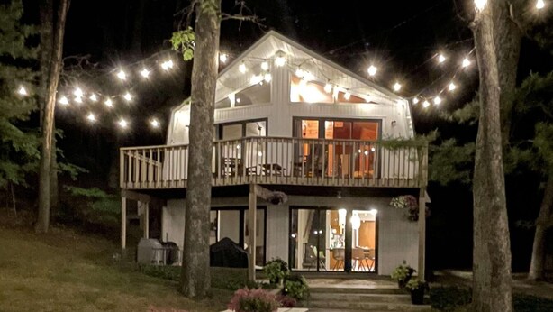 A two-story house with a balcony, string lights, and a picnic table with flowers. - Lakefront Cottage with Private Beach, Sleeps 14 (Newaygo)