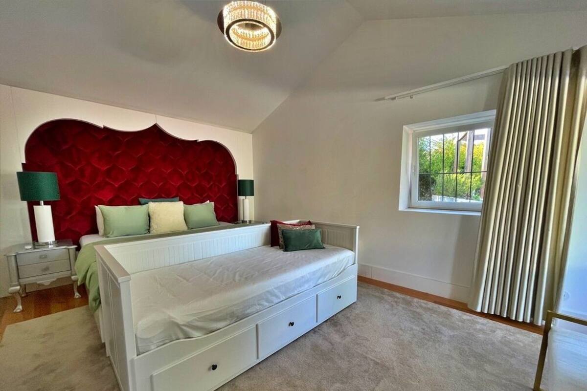 A bedroom with a large bed, a red upholstered headboard, a window with curtains, and a bedside table with lamps.