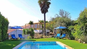 A swimming pool with blue lounge chairs and umbrellas, surrounded by greenery and a building with a red roof.