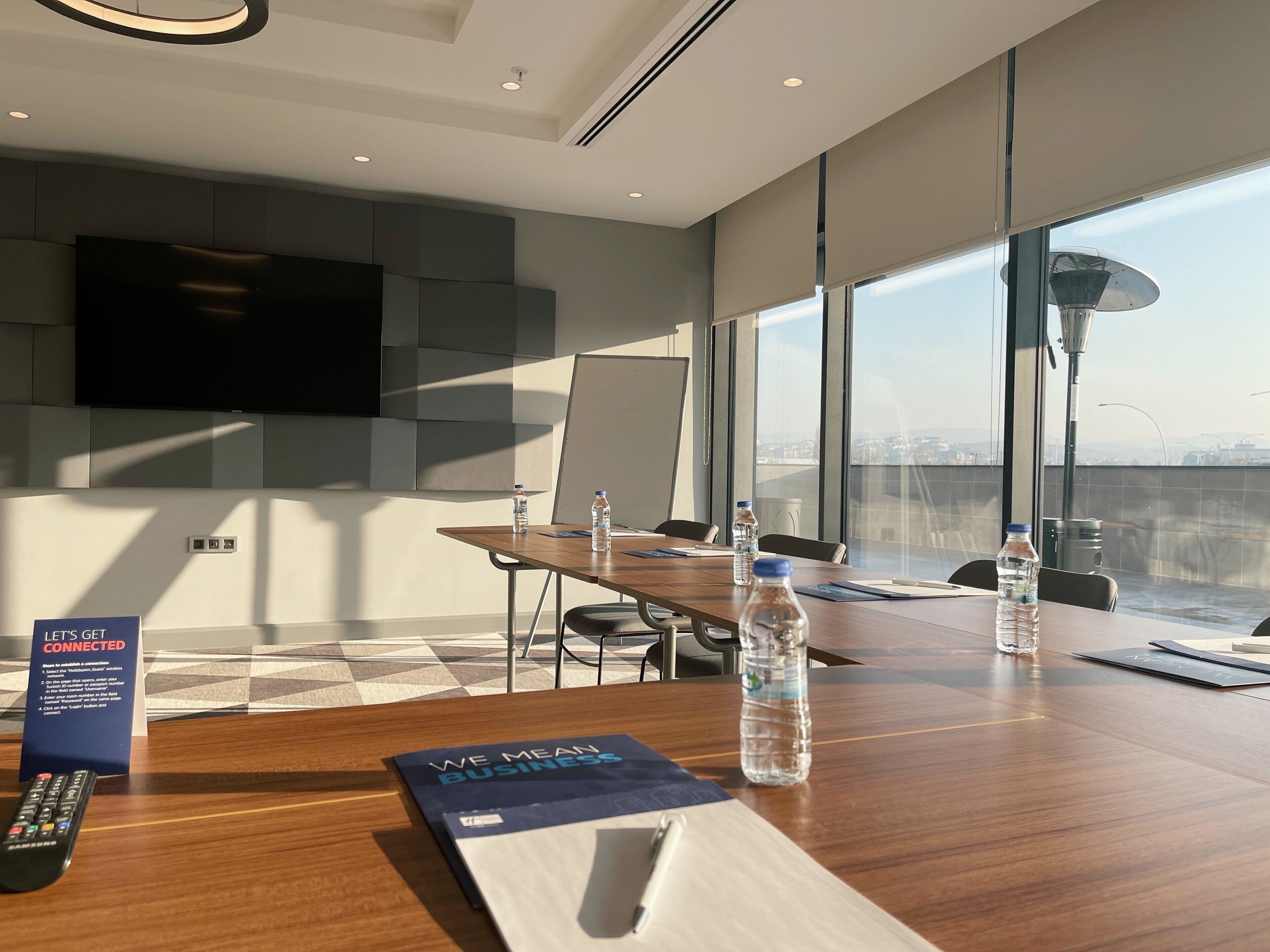 Una sala de conferencias moderna con una gran mesa, sillas, una pantalla de proyector y vistas de la ciudad a través de amplios ventanales.