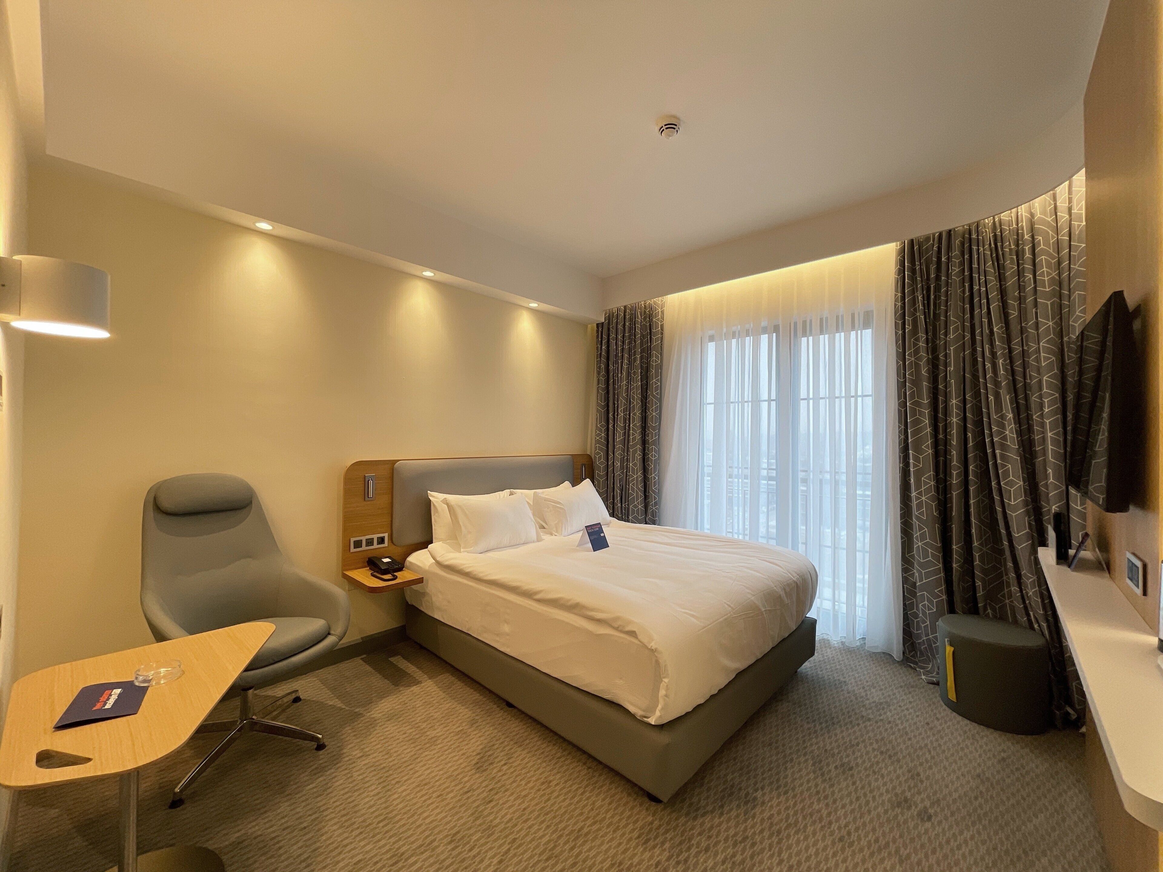 A hotel room with a bed, a desk, a chair, a TV, and a window with curtains.