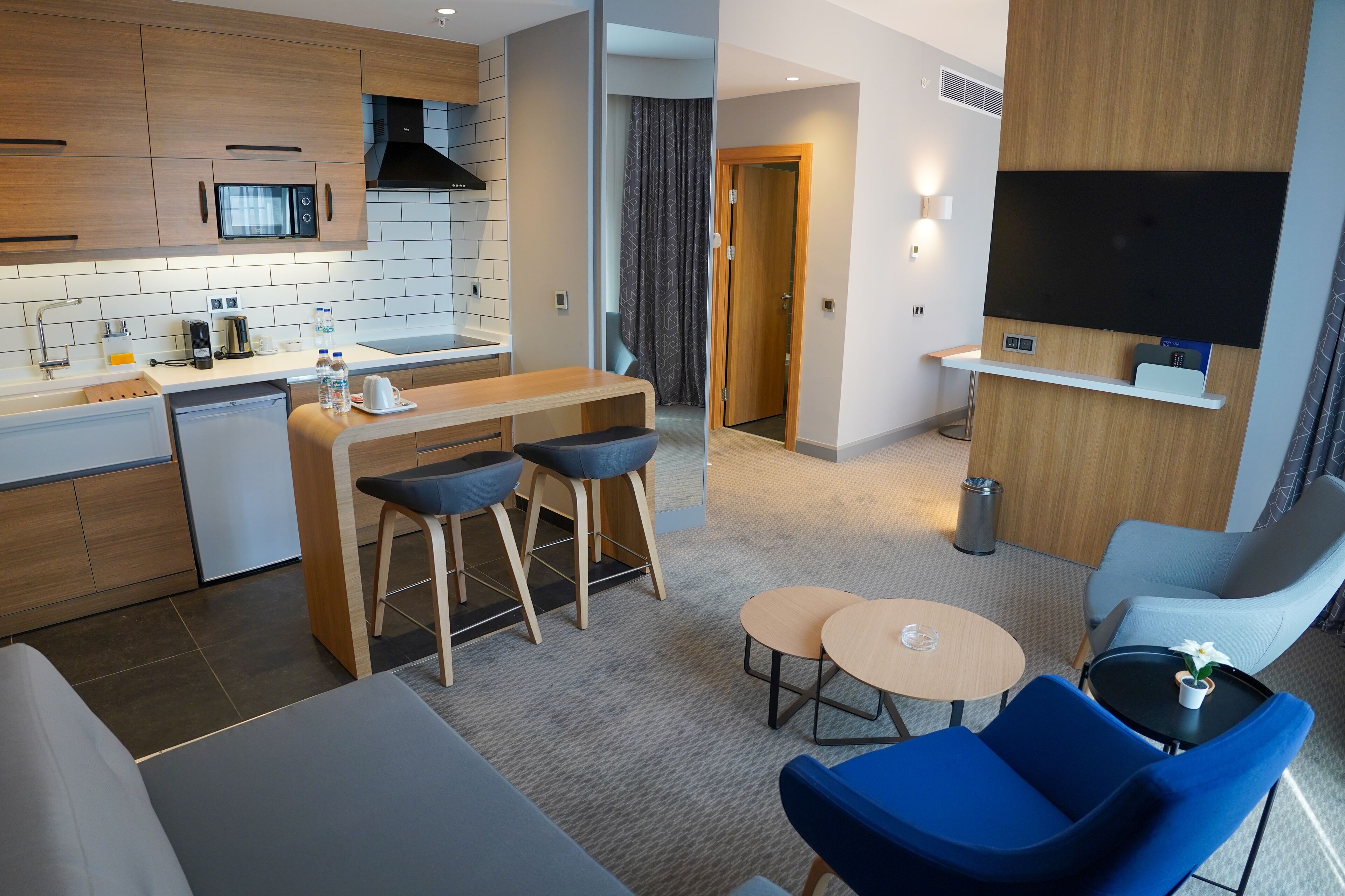 Una habitación de hotel moderna con kitchenette, un comedor con taburetes, un televisor y un área de descanso con sofá y sillones.