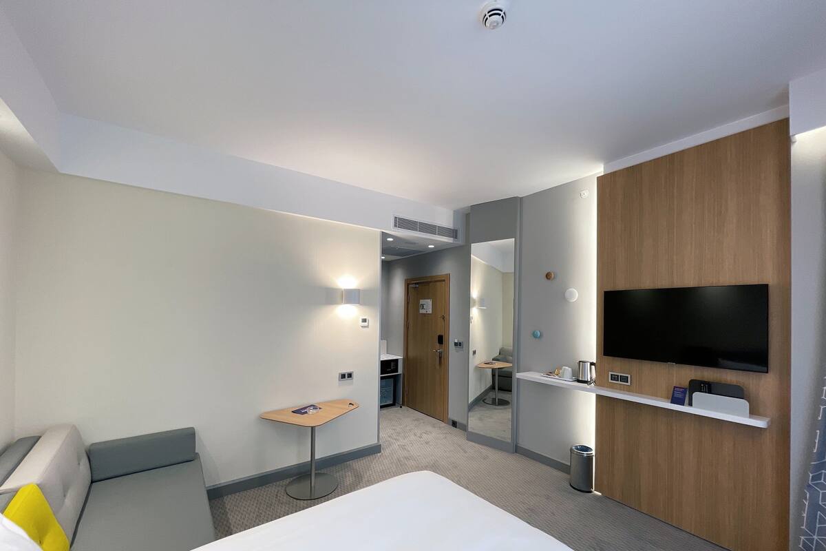 A modern hotel room with a bed, a small desk, a TV mounted on the wall, and a mirror.