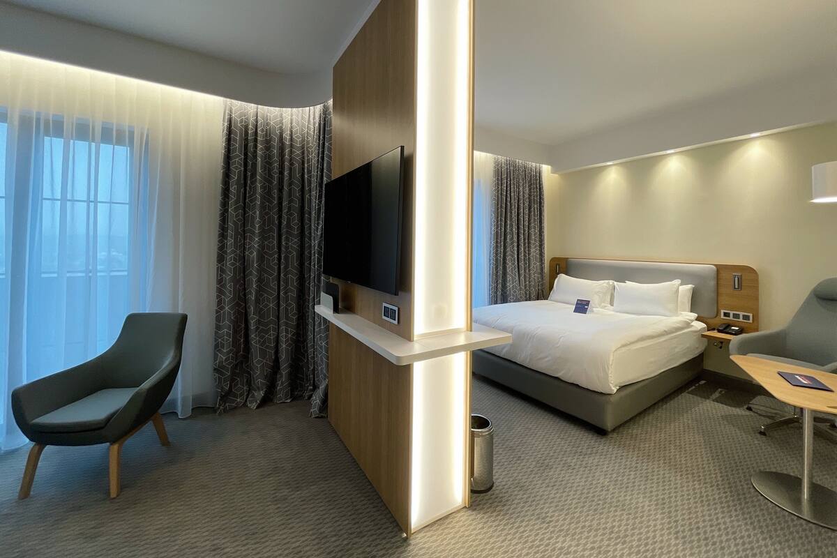 A modern hotel room with a bed, a TV mounted on a wooden panel, a desk with a chair, and a large window with curtains.