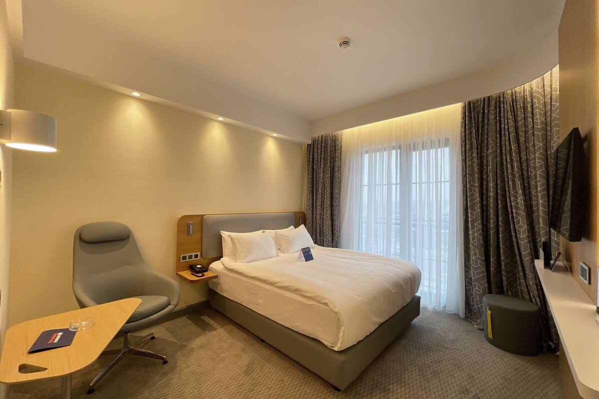 A hotel room with a bed, a desk, a chair, a TV, and a window with curtains.