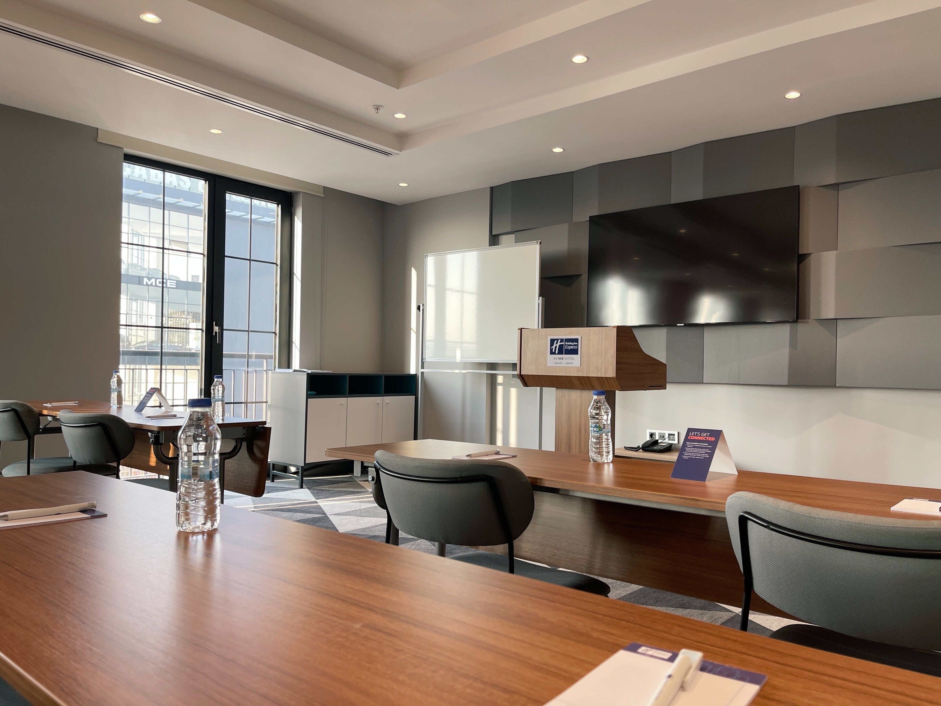 A modern hotel conference room with a wooden table, chairs, a large flat-screen TV, and a reception desk.