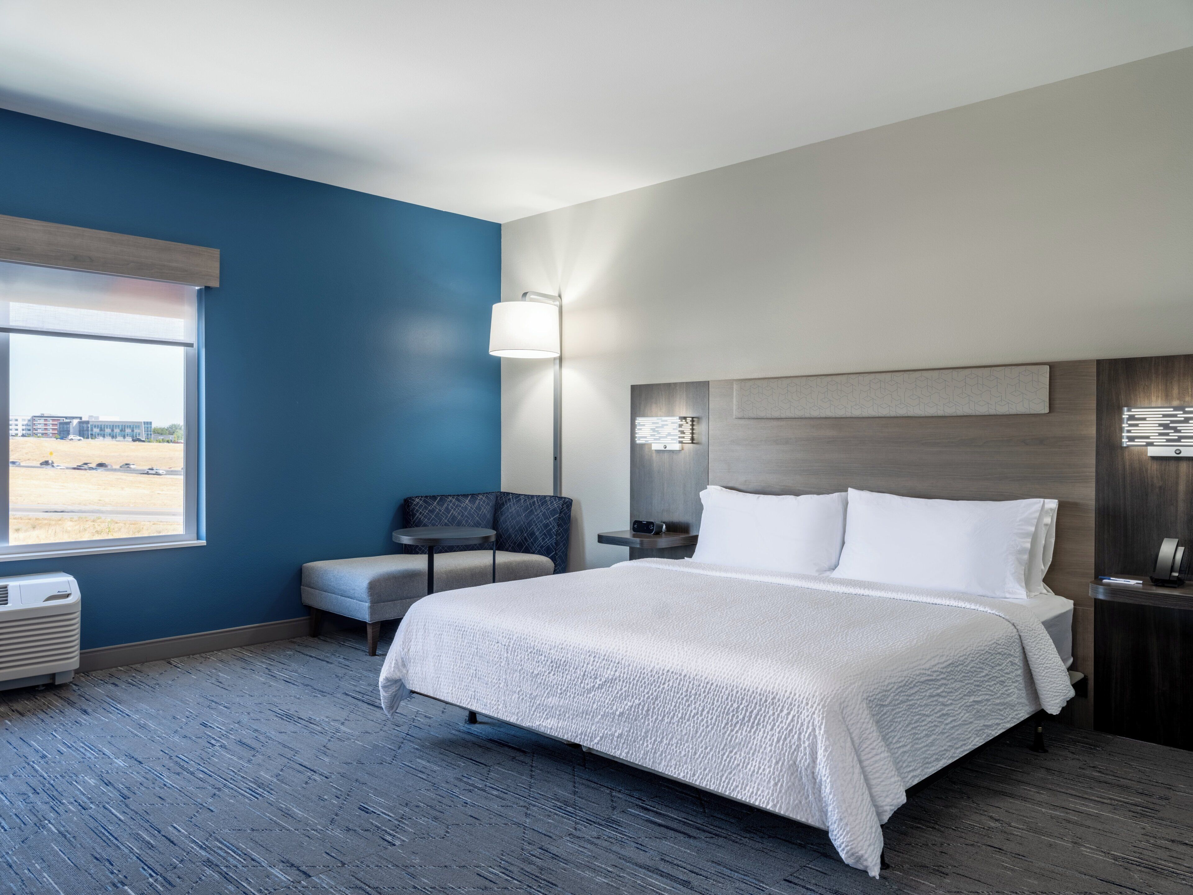 A hotel room with a large bed, a chair, a desk, and a window with a view of a building and parking lot.