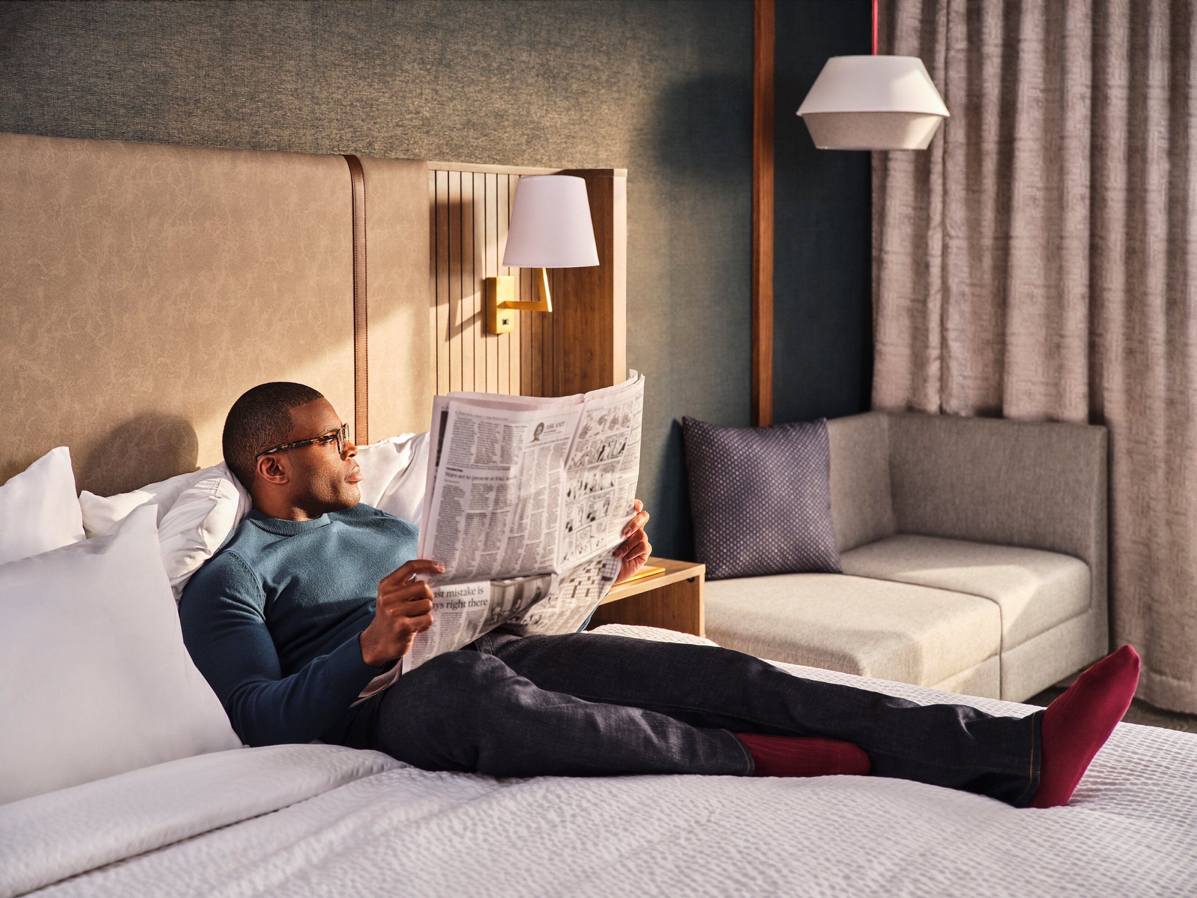 Una persona recostada en una cama leyendo un periódico en una habitación de hotel con cabecera de madera, una mesita de noche con lámpara y un sofá.
