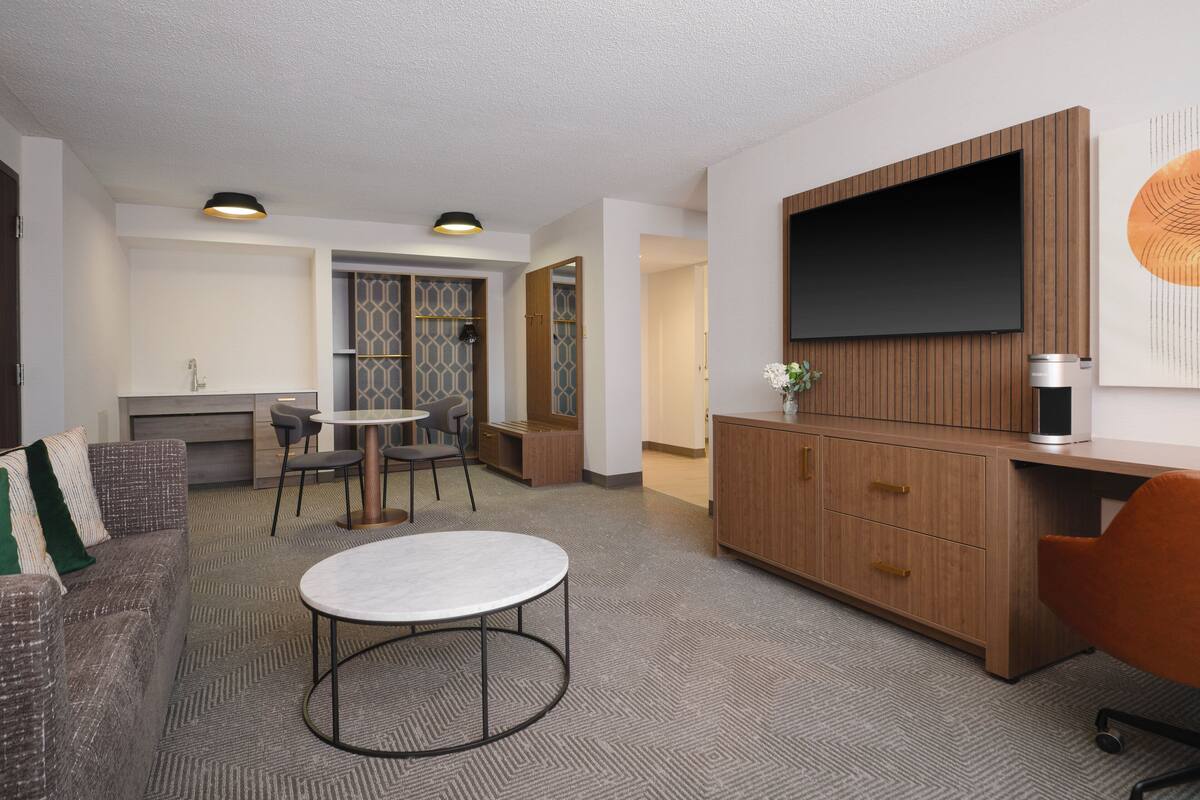 Ein modernes Wohnzimmer mit einer Couch, einem runden Couchtisch, einem Fernseher und einer Küchenzeile im Hintergrund.