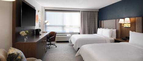 A hotel room with a desk, two beds, a chair, and a large window.