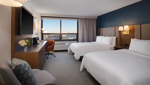 A hotel room with two beds, a desk, a chair, a TV, and a large window with a city view.