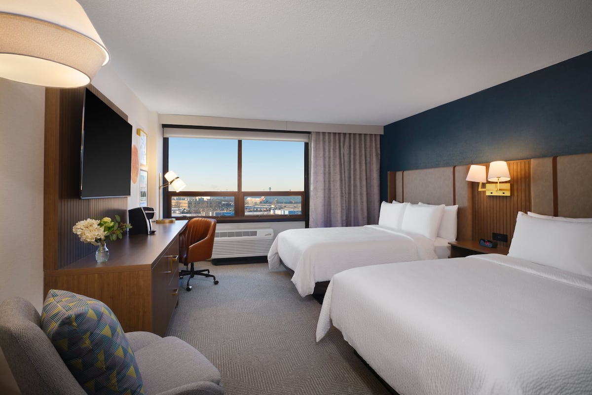 Ein Hotelzimmer mit zwei Betten, einem Schreibtisch, einem Stuhl, einem Fernseher und einem großen Fenster mit Blick auf die Stadt.
