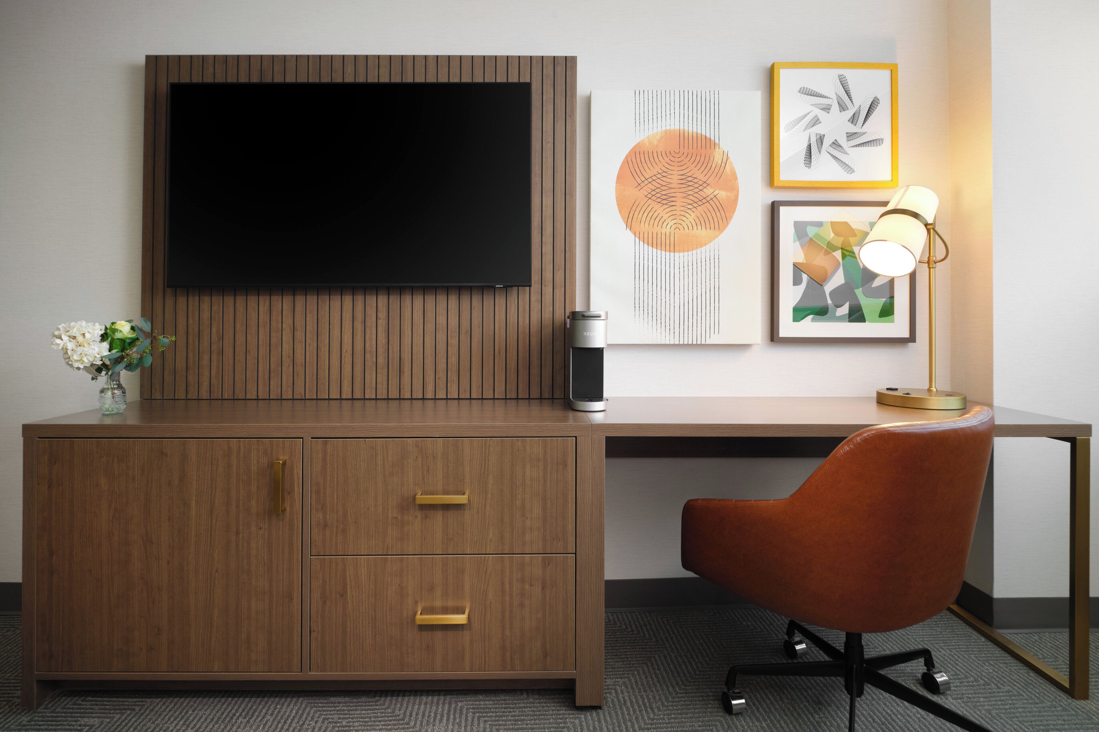 A modern hotel room with a flat-screen TV mounted on a wooden panel wall, a desk with a chair, and a coffee maker.