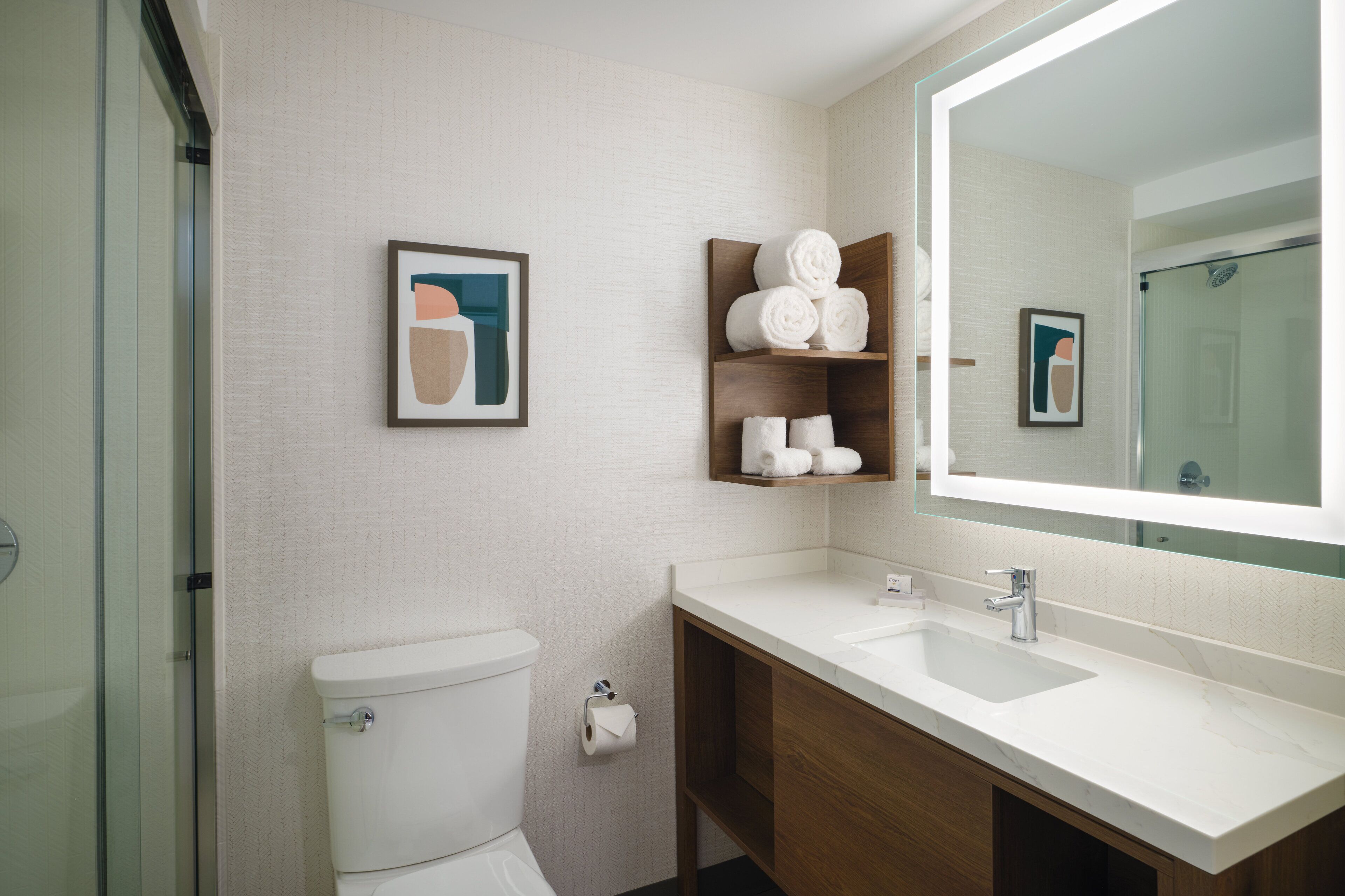 Et moderne badeværelse med toilet, vask og brusebad. Der er håndklæder og et billede i ramme på væggen.