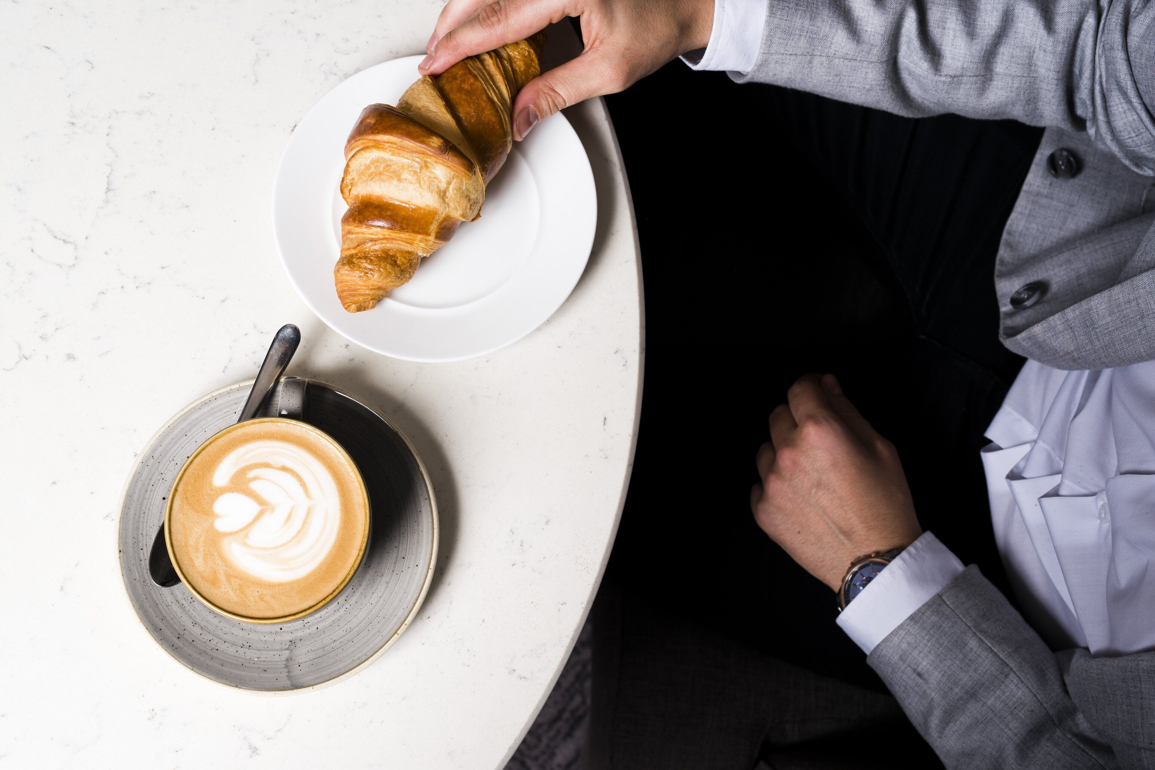 A person in a grey suit sitting at a table with a croissant and a cup of coffee.