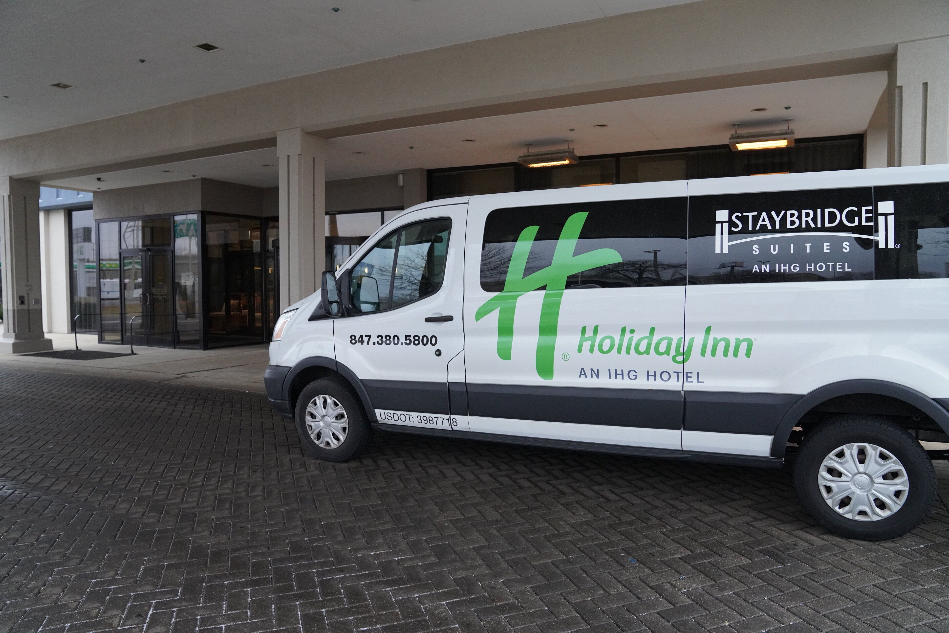 A white Holiday Inn Staybridge Suites van parked in front of a hotel entrance with a glass door and a brick pavement.