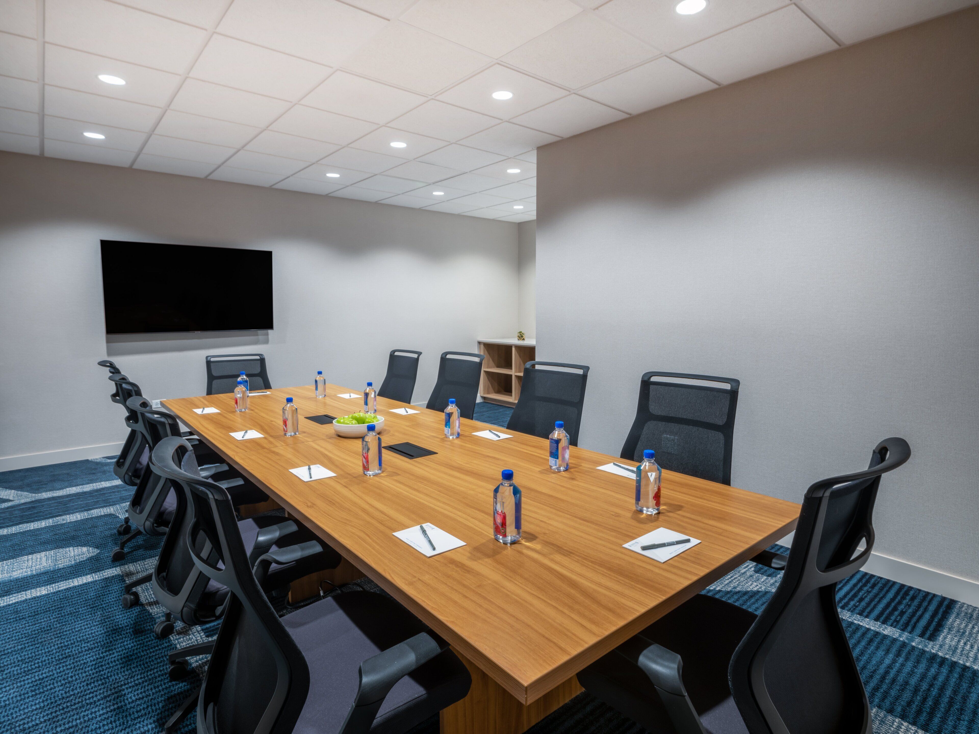 Un salón de conferencias con una larga mesa, sillas, botellas de agua y una pantalla.