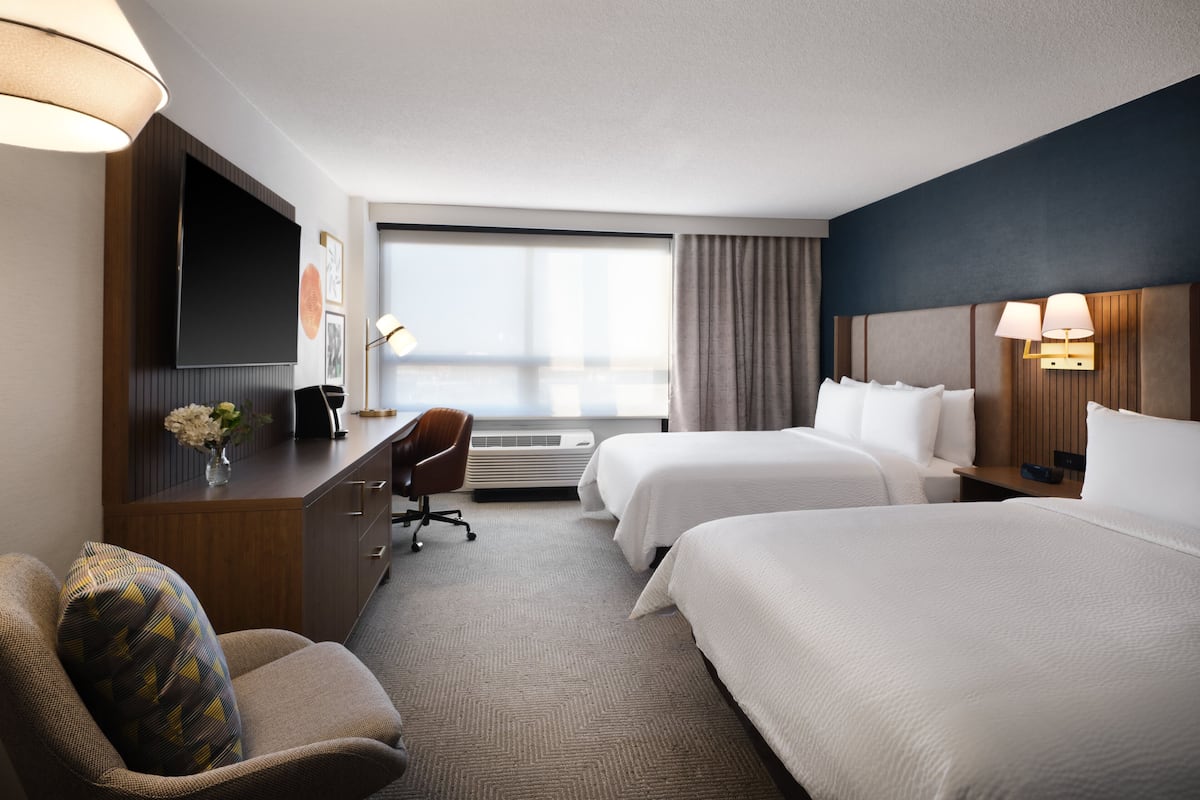 Ein Hotelzimmer mit zwei Betten, einem Schreibtisch, einem Stuhl, einem Fernseher und einem großen Fenster.