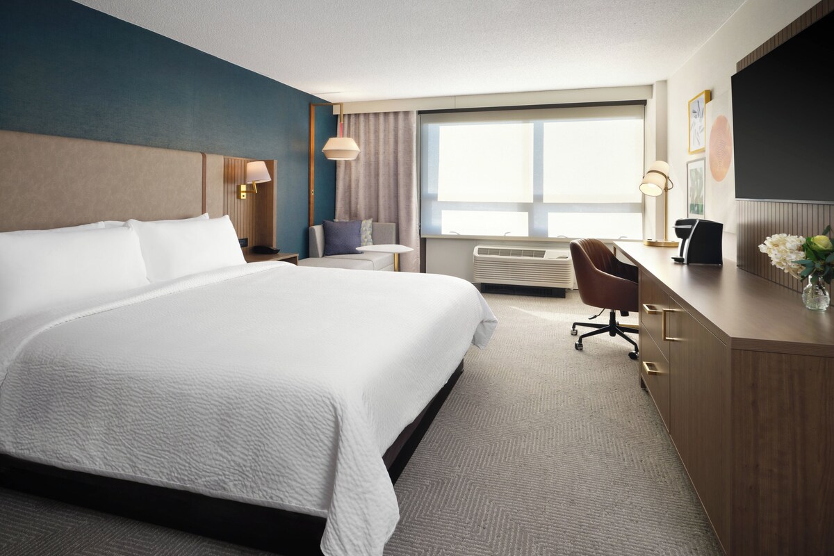 Een hotelkamer met een groot bed, een bureau met stoel, een klein tafeltje, een lamp en een groot raam met gordijnen.