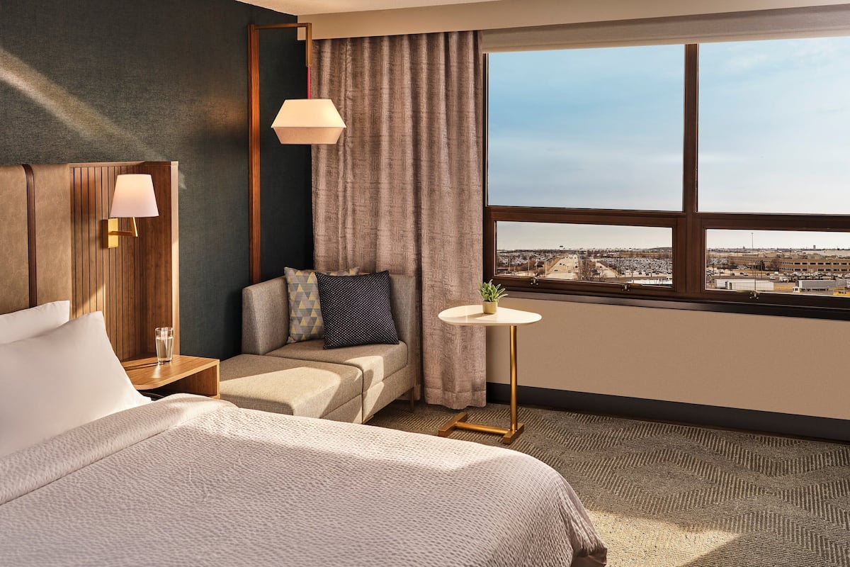 Ein Hotelzimmer mit einem großen Bett, einer kleinen Couch, einem Nachttisch mit Lampe, einem runden Tisch mit Pflanze und einem Fenster mit Blick auf die Stadt.