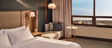 Una habitación de hotel con una cama grande, un pequeño sofá, una mesita de noche con lámpara, una mesa redonda con una planta y una ventana con vistas a la ciudad.