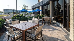 An outdoor patio with a wooden table and wicker chairs, surrounded by blue umbrellas, and a building with large windows in the background.