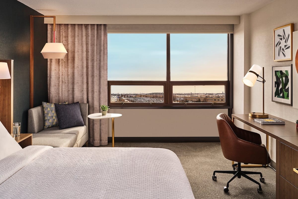 Ein Hotelzimmer mit einem großen Bett, einem Schreibtisch mit Stuhl, einem kleinen Tisch mit Pflanze, Blick auf die Stadt durch das Fenster und Kunstwerken an der Wand.