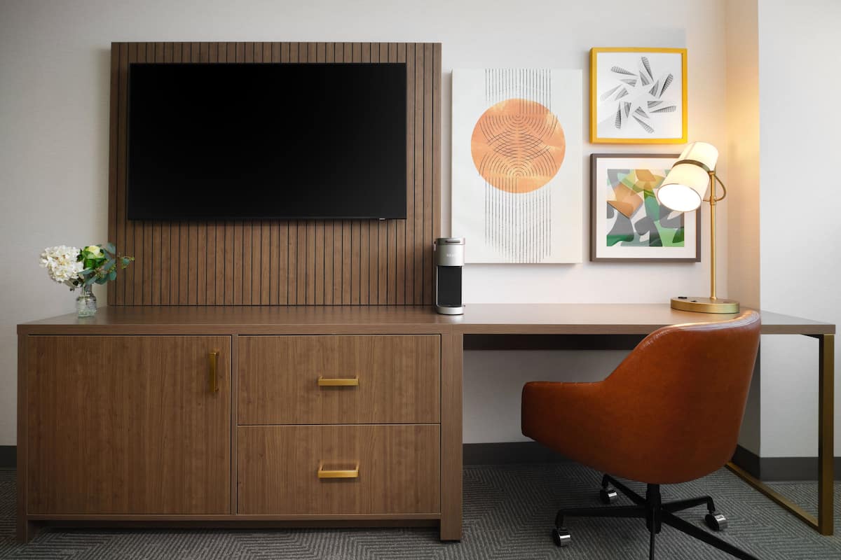 Ein modernes Hotelzimmer mit Schreibtisch aus Holz, Flachbildfernseher, Kaffeemaschine und bequemem Sessel.
