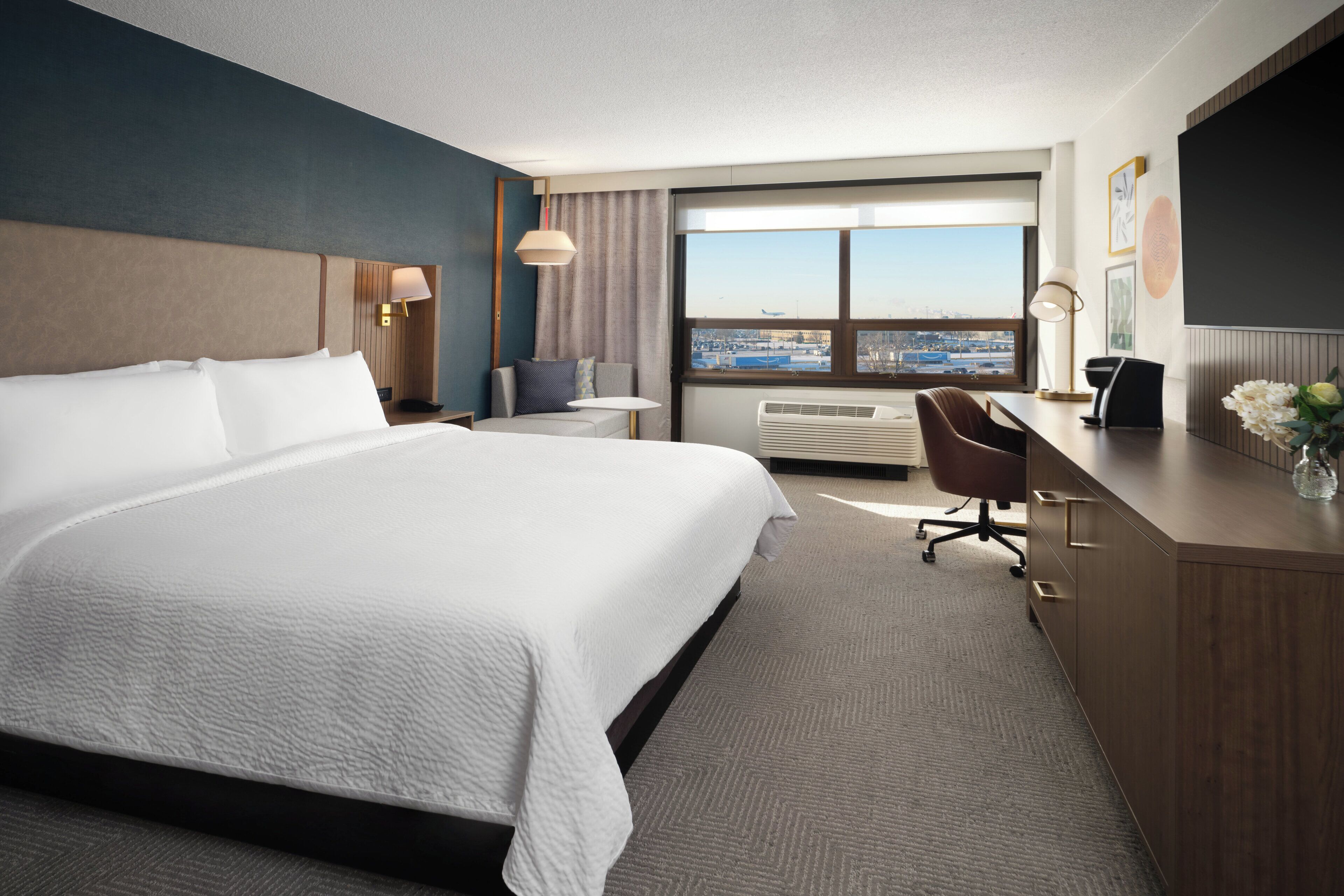 A hotel room with a large bed, a desk, a chair, a desk lamp, a flat-screen TV, and a large window with a city view.