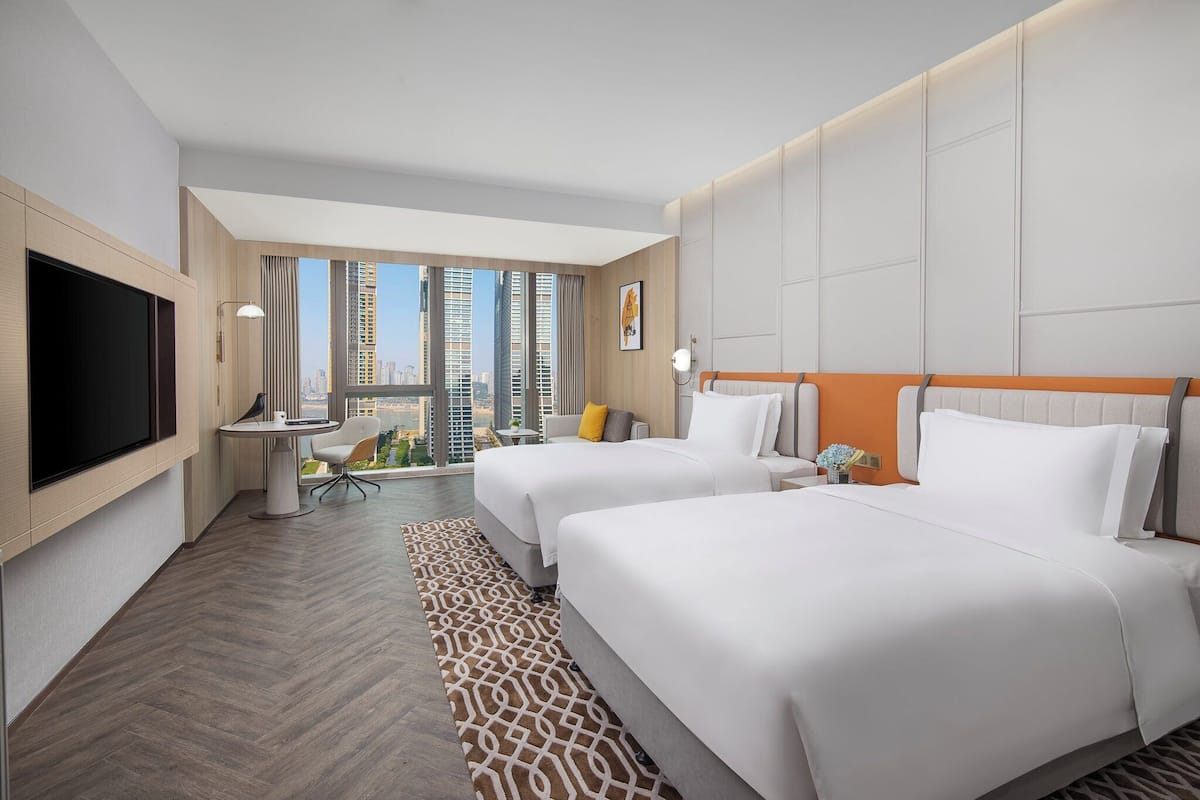 A hotel room with two beds, a TV, a desk, and a view of the city.