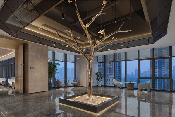 A modern lobby with a central tree sculpture, seating areas, and large windows offering city views.