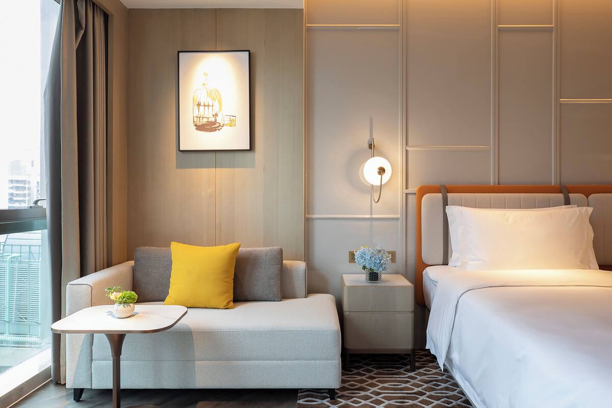 A hotel room with a sofa, a small table with a plant, a bedside table with a vase of flowers, a bed with white linens, and a framed picture on the wall.
