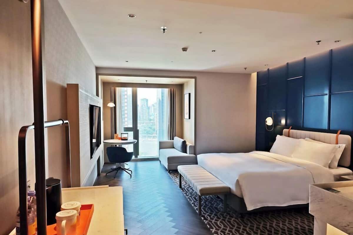 A modern hotel room with a large bed, a desk with a chair, a small table with two cups, and a view of the city through the window.