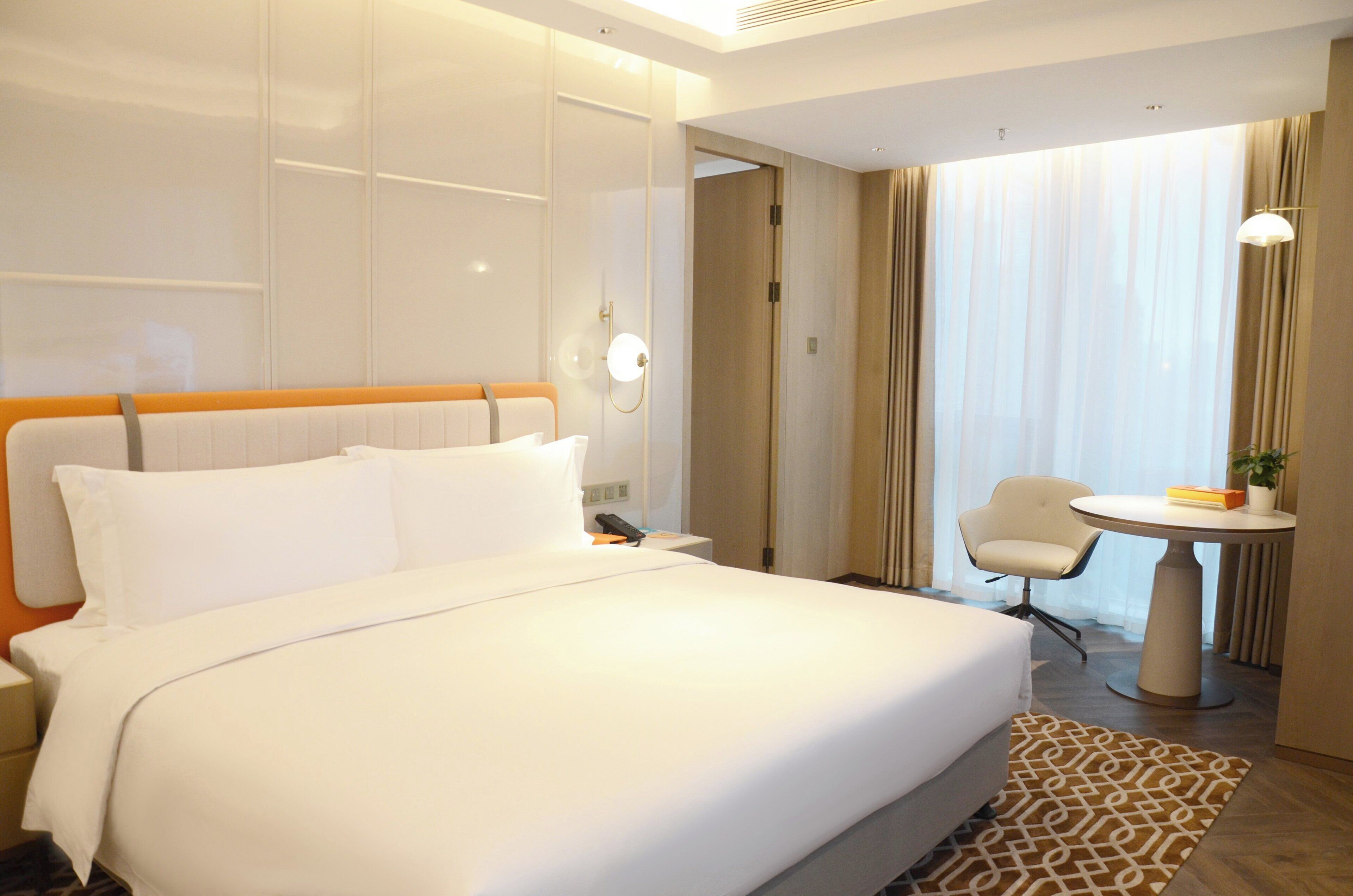 A hotel room with a large bed, a bedside table, a chair, a small table, and a window with curtains.
