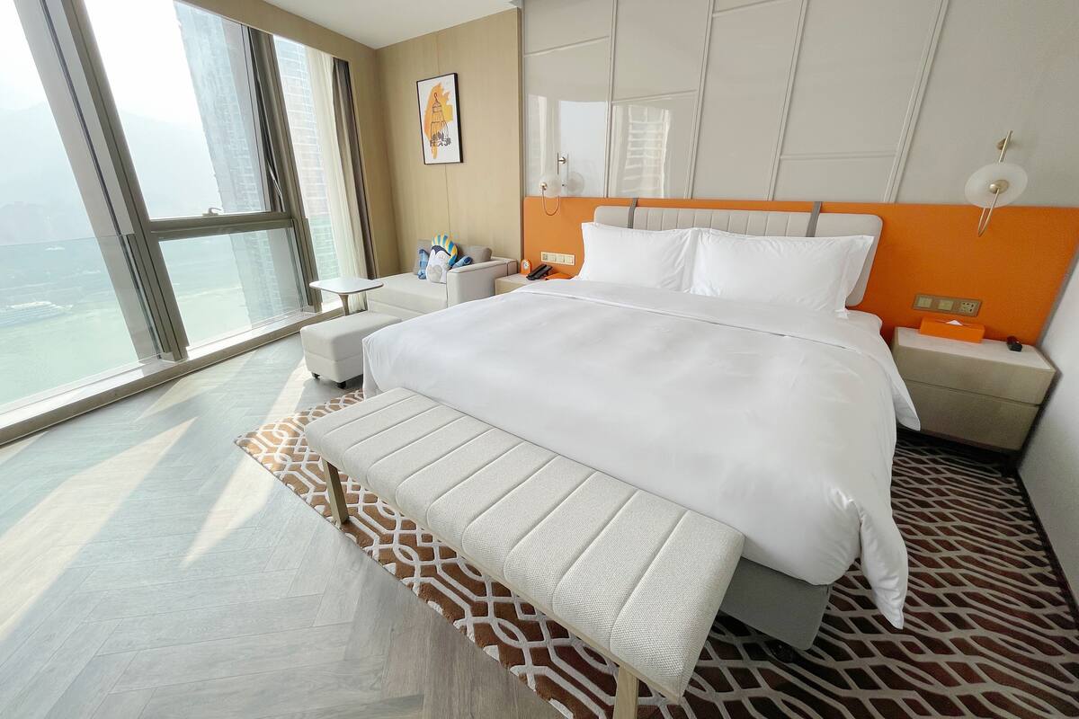 A modern hotel room with a large bed, a bedside table, a nightstand, a window with a view, and a patterned rug.